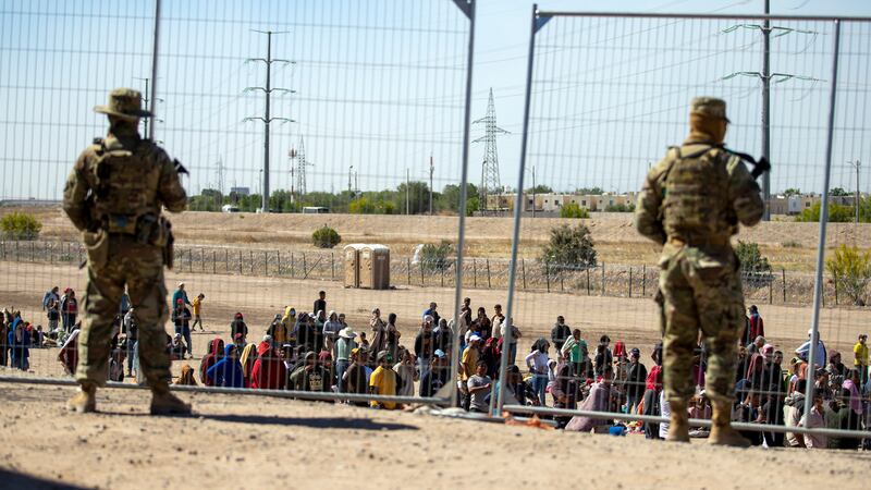FILE - Migrants wait in line adjacent to the border fence under the watch of the Texas...